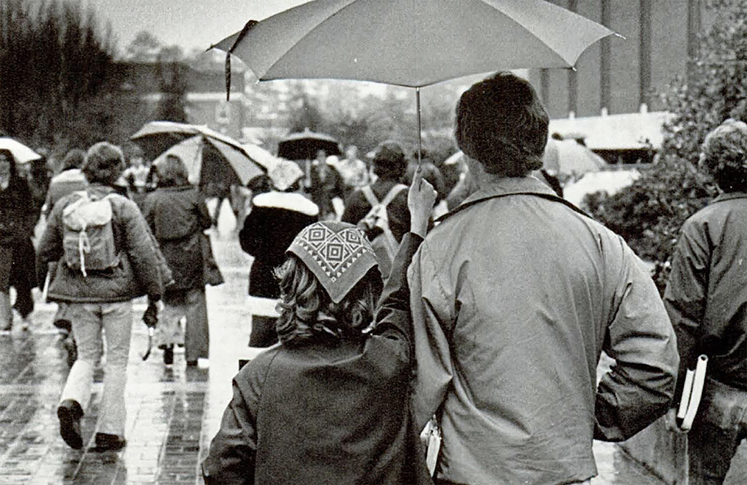 black and white photo of people walking in the ran on a university campus with the backs of young couple sharing an umbrella in focus